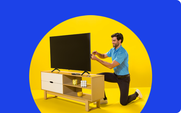 Un solucionador con una camisa azul claro está instalando un televisor de pantalla plana negro sobre un soporte de madera de color marrón claro contra un fondo amarillo brillante.