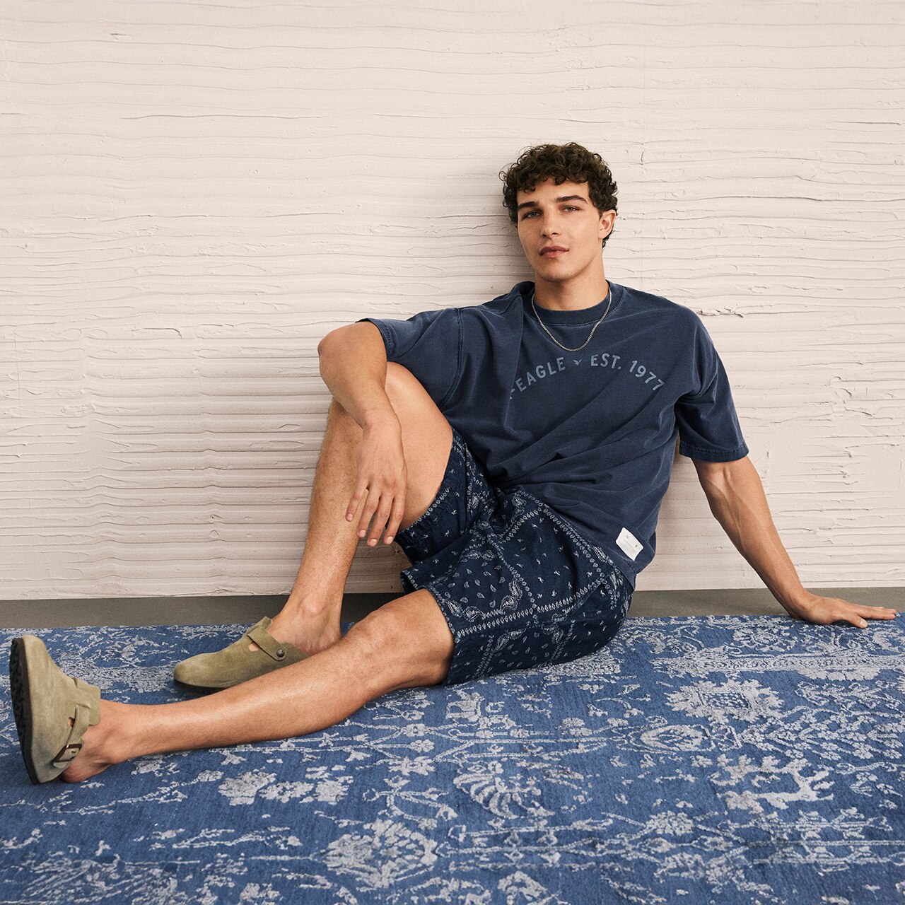 Modelo hombre usando playera azul con estampado, short azul y zapatos casuales verdes. Pulsa aquí para ver playeras gráficas de la marca American Eagle.
