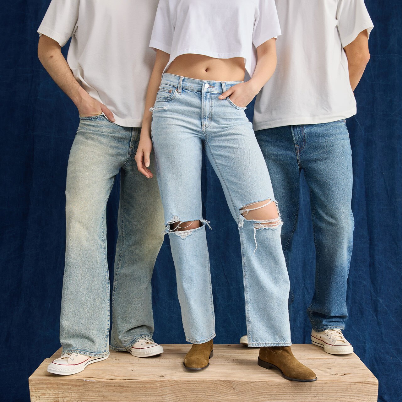 Dos modelos hombres y una modelo mujer usando playeras blancas y pantalones de mezclilla azul en diferentes modelos. Pulsa aquí para ver jeans de la marca American Eagle.