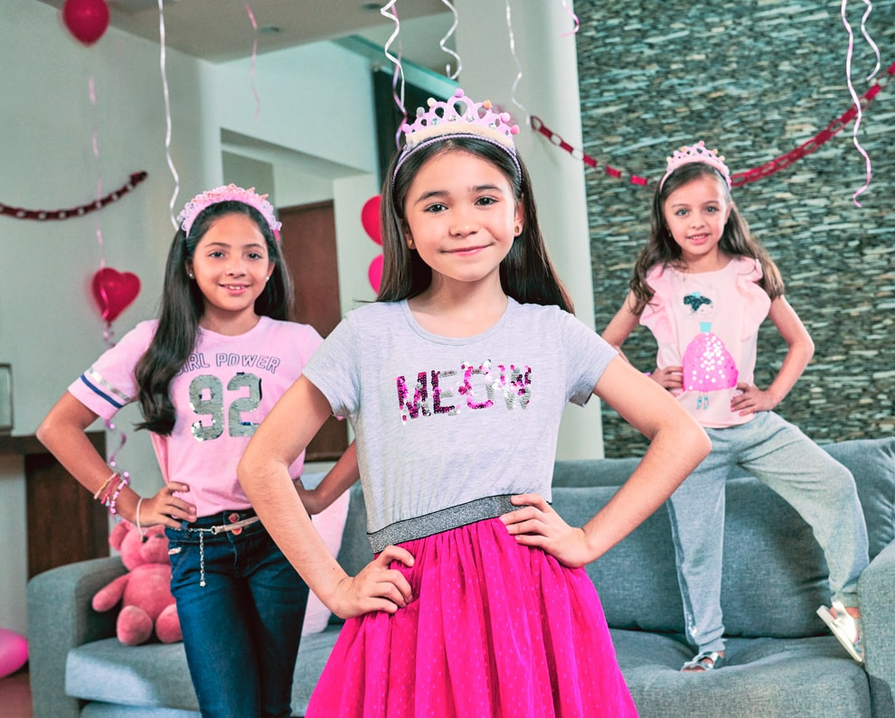 Tres niñas, una con playera rosa de manga corta con el número 92 gris al frente del torso y una corona rosa, otra con playera gris de manga corta con letras iridiscentes moradas y grises al frente del torso y una corona y otra con un vestido rosa sin mangas con una corona rosa. Pulsa aquí para encontrar más de Girls Attitude, abre en pestaña nueva.